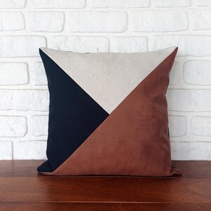 May include: Square decorative pillow with a geometric design. The pillow features a color block pattern with a navy blue, tan, and brown sections. The pillow is placed on a wooden surface in front of a white brick wall.