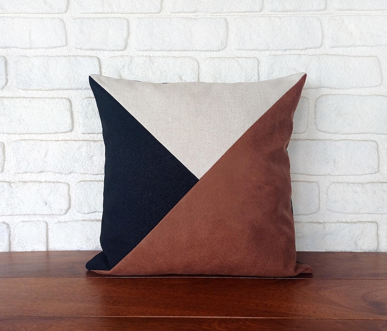 May include: Square decorative pillow with a geometric design. The pillow features a color block pattern with a navy blue, tan, and brown sections. The pillow is placed on a wooden surface in front of a white brick wall.