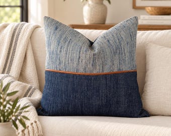 May include: A square throw pillow featuring a two-tone design with a light blue textured upper section and a dark navy blue textured lower section, separated by a thin, horizontal brown leather-like piping detail across the center.