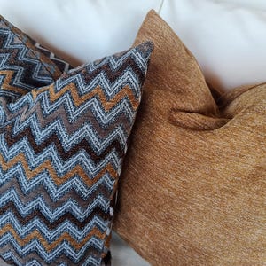 Op de afbeelding: Drie decoratieve kussens. E&eacute;n kussen heeft een bruin, blauw en goud zigzagpatroon. Het tweede kussen is effen, getextureerd en goudbruin van kleur. De kussens liggen tegen een witte achtergrond.
