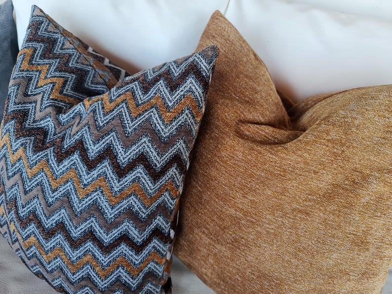 Op de afbeelding: Drie decoratieve kussens. E&eacute;n kussen heeft een bruin, blauw en goud zigzagpatroon. Het tweede kussen is effen, getextureerd en goudbruin van kleur. De kussens liggen tegen een witte achtergrond.