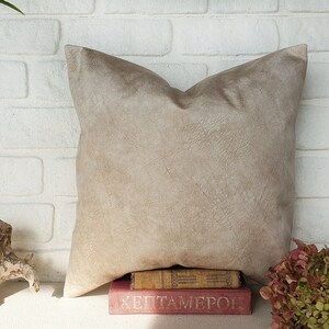 May include: A beige suede pillow with a subtle textured surface, resting on a stack of books with the title "Heptameron" visible. The pillow is in front of a white brick wall.