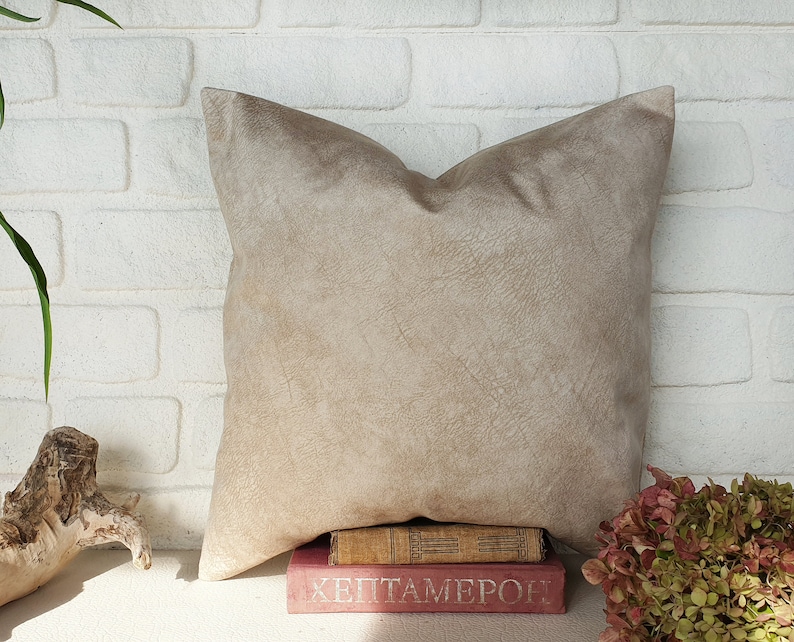 May include: A beige suede pillow with a subtle textured surface, resting on a stack of books with the title "Heptameron" visible. The pillow is in front of a white brick wall.