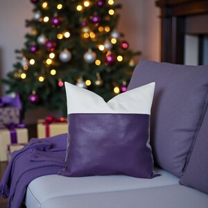 May include: A decorative pillow with a white top and a deep purple bottom rests on a gray couch. A purple throw blanket and other purple pillows are also on the couch. A Christmas tree with purple and silver ornaments is in the background.
