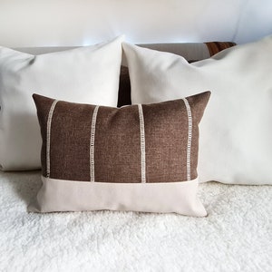 May include: A brown and white striped throw pillow with a white leather bottom. The pillow is sitting on a white fluffy bedspread.