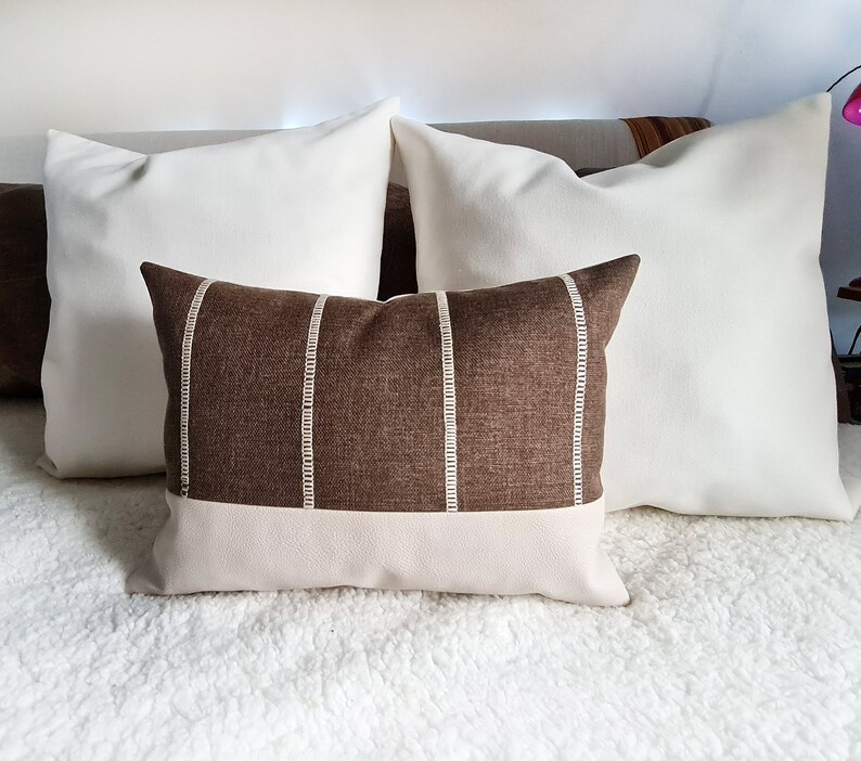 May include: A brown and white striped throw pillow with a white leather bottom. The pillow is sitting on a white fluffy bedspread.