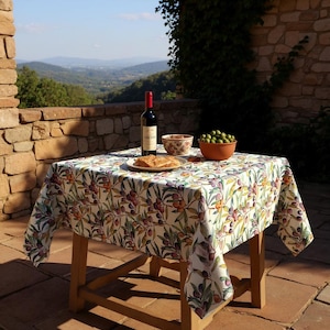 Pode incluir: Uma mesa de madeira coberta com uma toalha de mesa com um padrão colorido de ramos de oliveira. Uma garrafa de vinho tinto, uma tigela de azeitonas, uma tigela de nozes e um pão estão sobre a mesa. A cena é ao ar livre com vista para a montanha.