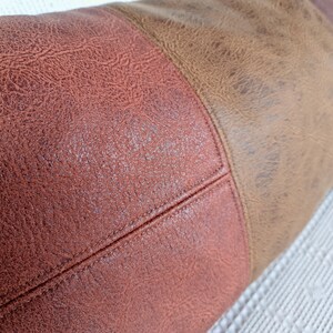 May include: A cylindrical bolster pillow featuring a two-tone design with a textured reddish-brown section joined to a lighter tan section by a visible stitched seam. The surface has a distinct pebbled grain pattern throughout the fabric.