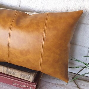 Puede incluir: Una almohada de cuero marr&oacute;n con un acabado texturizado sutil. La almohada tiene una forma rectangular con costuras cosidas y un borde ligeramente redondeado.