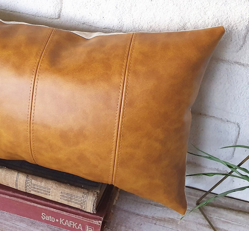 Puede incluir: Una almohada de cuero marr&oacute;n con un acabado texturizado sutil. La almohada tiene una forma rectangular con costuras cosidas y un borde ligeramente redondeado.