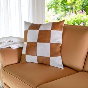 May include: A brown and white checkered throw pillow with a faux leather design. The pillow is sitting on a brown couch.