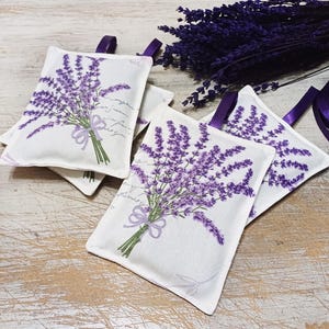 Op de afbeelding: Vier witte stoffen lavendelzakjes met bedrukte paarse lavendelbloemen. De zakjes hebben een lint aan de bovenkant om ze op te hangen.