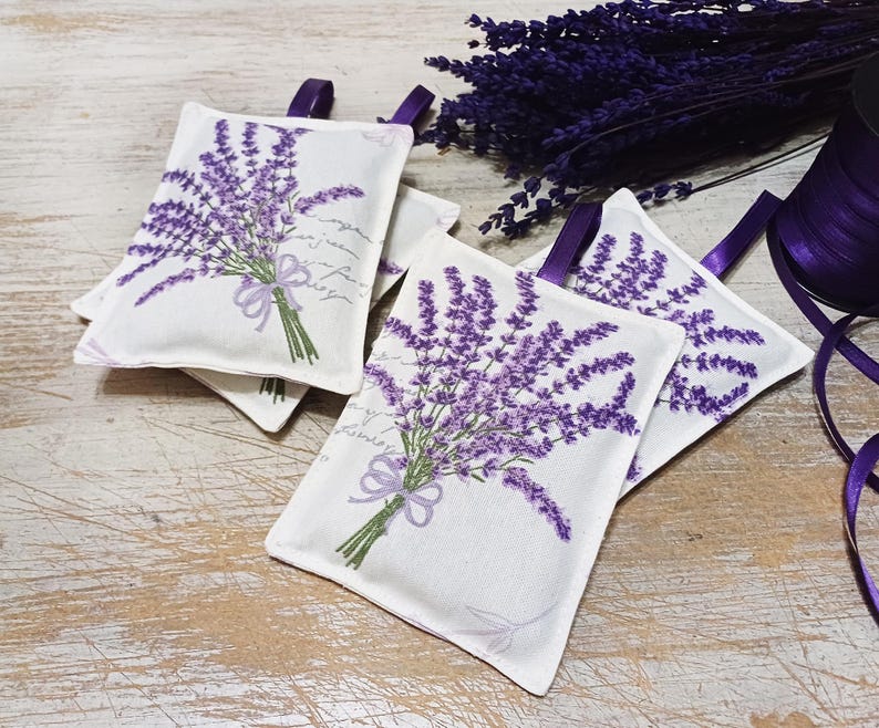 Op de afbeelding: Vier witte stoffen lavendelzakjes met bedrukte paarse lavendelbloemen. De zakjes hebben een lint aan de bovenkant om ze op te hangen.