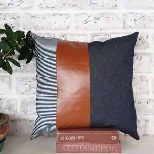 Puede incluir: Una almohada decorativa con una tela a rayas azul y blanca, un panel de cuero marrón y una tela vaquera azul oscuro. La almohada está encima de dos libros.