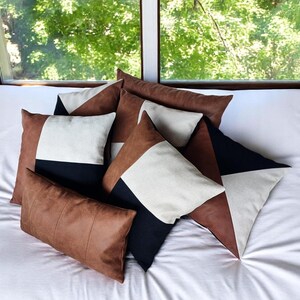 May include: A collection of decorative throw pillows in various shapes and sizes. The pillows feature geometric designs with brown, black, and cream-colored panels. The pillows are arranged on a white bedspread, with a window and greenery in the background.