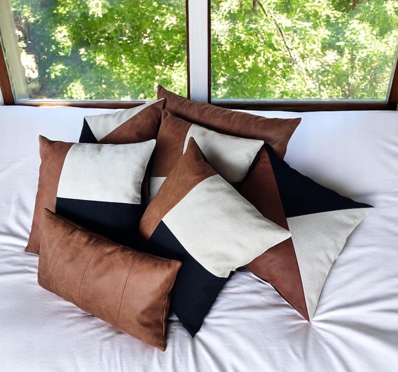 May include: A collection of decorative throw pillows in various shapes and sizes. The pillows feature geometric designs with brown, black, and cream-colored panels. The pillows are arranged on a white bedspread, with a window and greenery in the background.