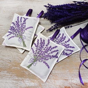 Op de afbeelding: Vier lavendelgeurzakjes met een witte achtergrond en bedrukte paarse lavendelbloemen. De zakjes zijn gebonden met een paars lint en hebben een handgeschreven bericht.