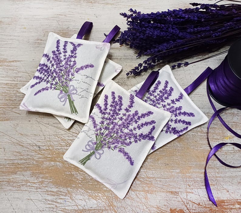 Op de afbeelding: Vier lavendelgeurzakjes met een witte achtergrond en bedrukte paarse lavendelbloemen. De zakjes zijn gebonden met een paars lint en hebben een handgeschreven bericht.