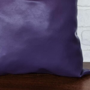 May include: A purple pillow with a smooth, leather-like texture. The pillow is sitting on a wooden surface.