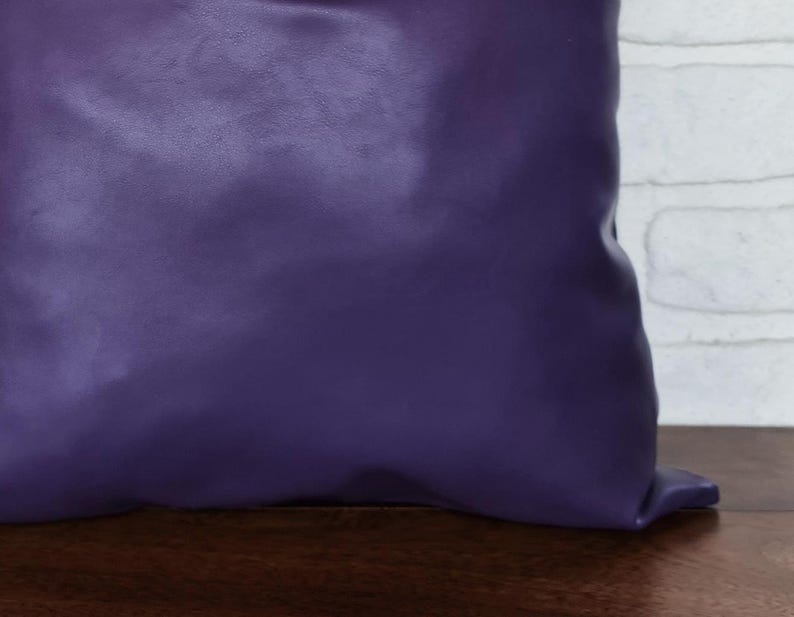 May include: A purple pillow with a smooth, leather-like texture. The pillow is sitting on a wooden surface.