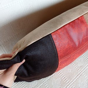 May include: A rectangular throw cushion featuring three distinct vertical panels of textured faux leather material in beige, dark brown, and a vibrant reddish-orange colour, joined together with visible stitching along the seams.