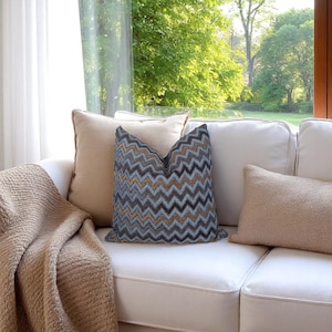Op de afbeelding: Een witte bank met diverse sierkussens en een plaid. De kussens omvatten een blauw, grijs en goud chevron patroon kussen, en twee effen beige kussens. Een beige plaid is over de bank gedrapeerd.
