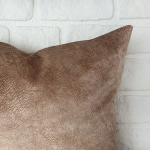 May include: A brown suede pillow with a textured surface. The pillow is shown against a white brick wall.