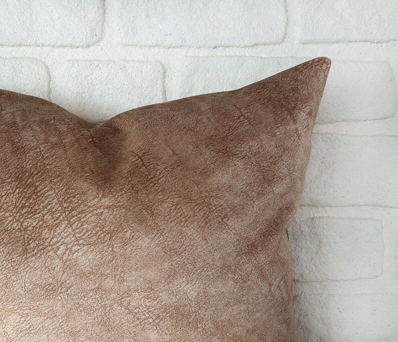 May include: A brown suede pillow with a textured surface. The pillow is shown against a white brick wall.