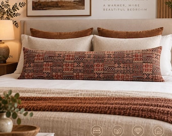 May include: An extra long lumbar pillow featuring a rectangular patchwork pattern in shades of rust, brown, and black. The pillow is positioned on a bed with white bedding, layered behind two solid rust-colored pillows and two beige pillows.