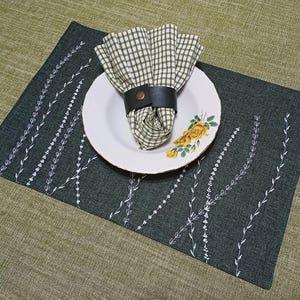 May include: A dark green placemat with a white embroidered design of leaves. A white plate with a yellow floral pattern sits on top of the placemat. A checkered green and white napkin is on the plate, with a brown leather napkin ring.