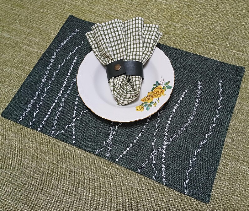 May include: A dark green placemat with a white embroidered design of leaves. A white plate with a yellow floral pattern sits on top of the placemat. A checkered green and white napkin is on the plate, with a brown leather napkin ring.