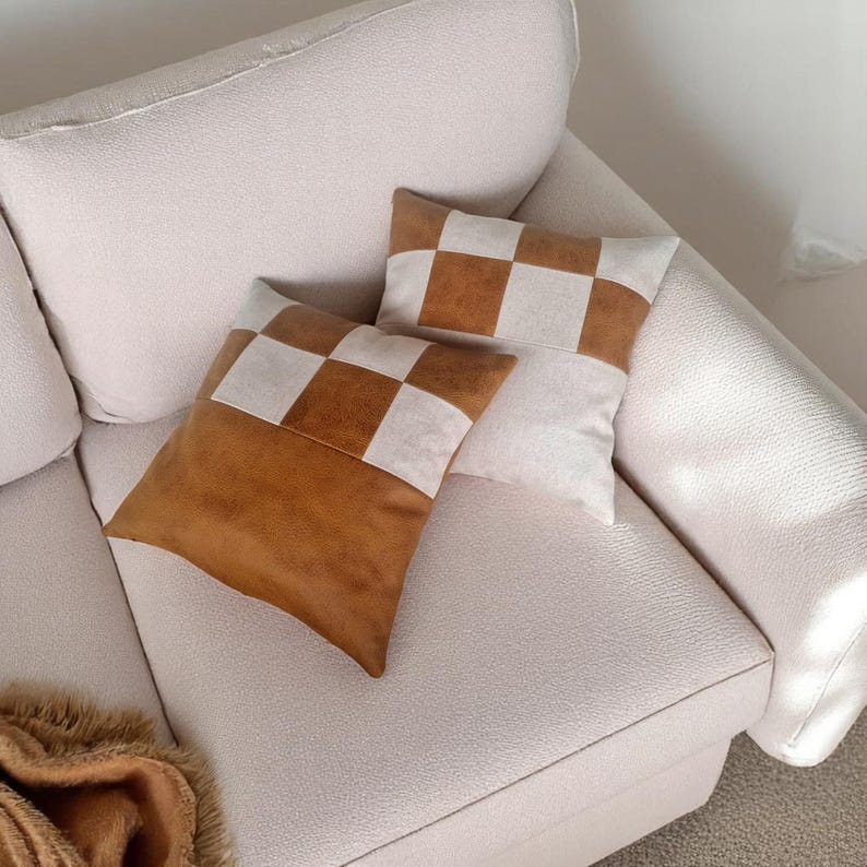 May include: Two decorative throw pillows with a checkered pattern. The pillows are made of a brown faux leather and white fabric. The pillows are on a white couch.