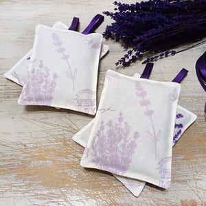 Op de afbeelding: Vier lavendelgeurzakjes met een bloemenmotief en de tekst "Fleur de Provence". De zakjes zijn gemaakt van witte stof met paarse lavendelbloemen en vlinders.