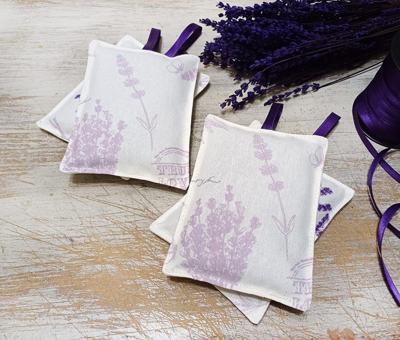 Op de afbeelding: Vier lavendelgeurzakjes met een bloemenmotief en de tekst "Fleur de Provence". De zakjes zijn gemaakt van witte stof met paarse lavendelbloemen en vlinders.