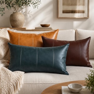 May include: Four leather throw pillows in shades of brown and teal rest on a cream-colored sofa. The pillows have vertical stitched detailing. A tan throw blanket with fringe is draped over the sofa arm. A vase with greenery is in the background.