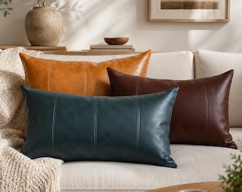 May include: Four leather throw pillows in shades of brown and teal rest on a cream-colored sofa. The pillows have vertical stitched detailing. A tan throw blanket with fringe is draped over the sofa arm. A vase with greenery is in the background.