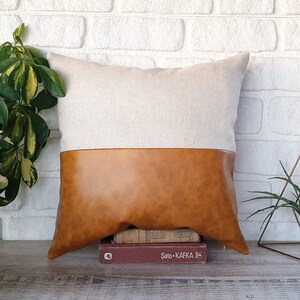 Puede incluir: Una almohada de lino beige con la parte inferior de piel sintética marrón. La almohada está sobre una superficie de madera con una pila de libros y un soporte de plantas de metal dorado.