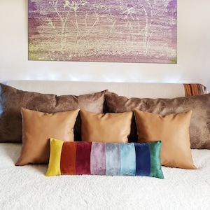 May include: A white bed with a brown faux leather headboard and a white fluffy blanket. There are five pillows on the bed, three brown faux leather pillows, two tan faux leather pillows, and a long rainbow velvet pillow.