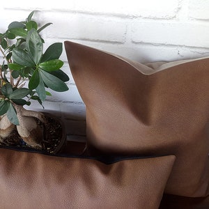 May include: Two brown faux leather throw pillows. The larger pillow has a light brown backing. The smaller pillow has a black trim.