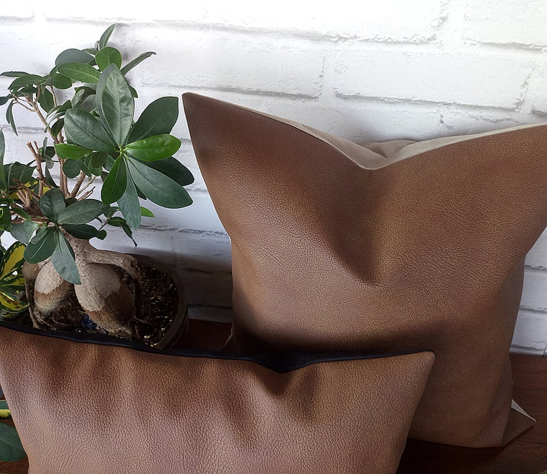 May include: Two brown faux leather throw pillows. The larger pillow has a light brown backing. The smaller pillow has a black trim.
