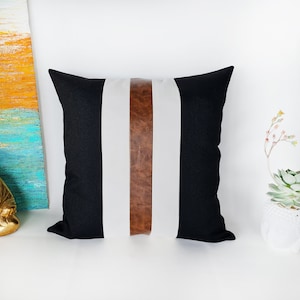 May include: Black and white striped cushion with a brown leather accent strip. The cushion is made of a soft, comfortable fabric and has a simple, modern design.
