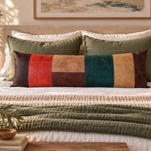 May include: A rectangular lumbar cushion featuring a patchwork design of leather-like panels in dark brown, bright red, tan, and deep teal. The cushion is positioned horizontally on a bed in front of two solid olive green rectangular cushions.