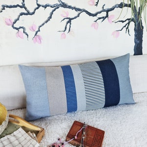 May include: A long rectangular pillow with a patchwork design featuring stripes of blue, white, and gray fabric. The pillow is lying on a white fluffy surface.