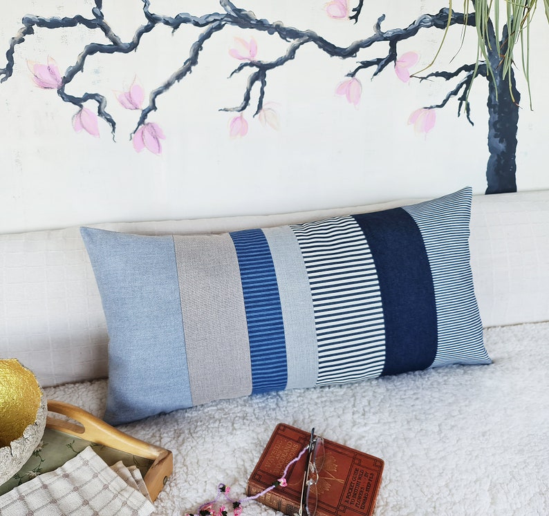May include: A long rectangular pillow with a patchwork design featuring stripes of blue, white, and gray fabric. The pillow is lying on a white fluffy surface.