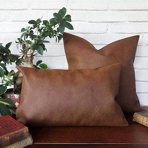 May include: Two brown leather throw pillows on a wooden surface with a potted plant in the background.