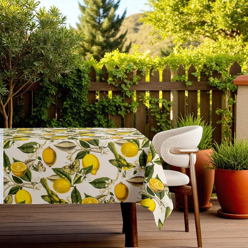 May include: A rectangular tablecloth with a lemon and bird pattern. The tablecloth is white with yellow lemons, green leaves, and small birds. The tablecloth is on a wooden table, with a white chair and potted plants in the background.