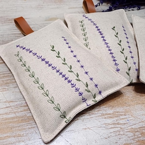 May include: Three beige linen sachets with brown leather ties. Each sachet features an embroidered design of purple lavender flowers and green leaves.