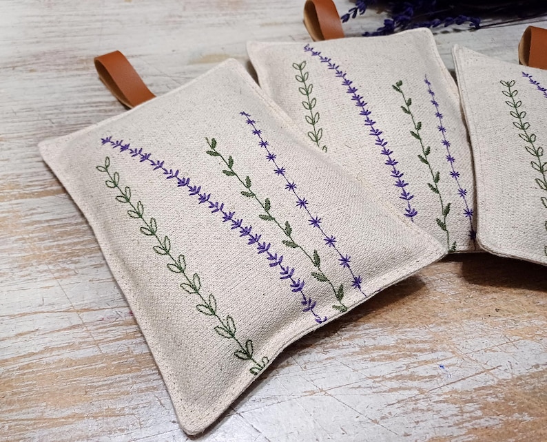 May include: Three beige linen sachets with brown leather ties. Each sachet features an embroidered design of purple lavender flowers and green leaves.