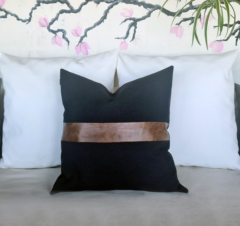 Puede incluir: Una almohada negra con una banda de cuero marr&oacute;n en el centro. La almohada est&aacute; sentada en un sof&aacute; blanco con dos almohadas blancas detr&aacute;s.