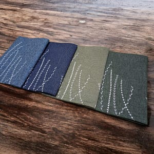 May include: Four fabric placemats with a white embroidered design of a plant with leaves. The placemats are in shades of blue and green.